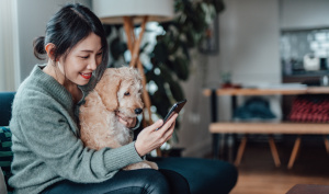 Asian woman sitting on couch with dog while looking at mobile phone